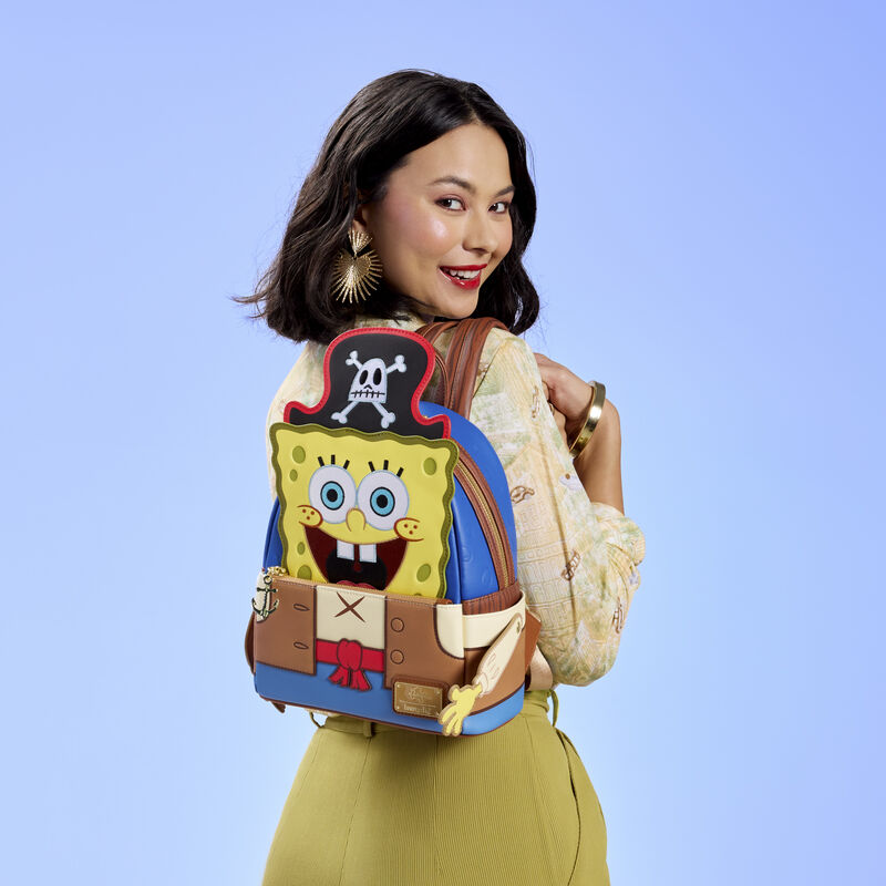 Woman facing away from camera wearing a light yellow blouse and olive green skirt. She wears the Loungefly SpongeBob SquarePants Pirate Costume Mini Backpack over a shoulder. She stands against a blue background. 